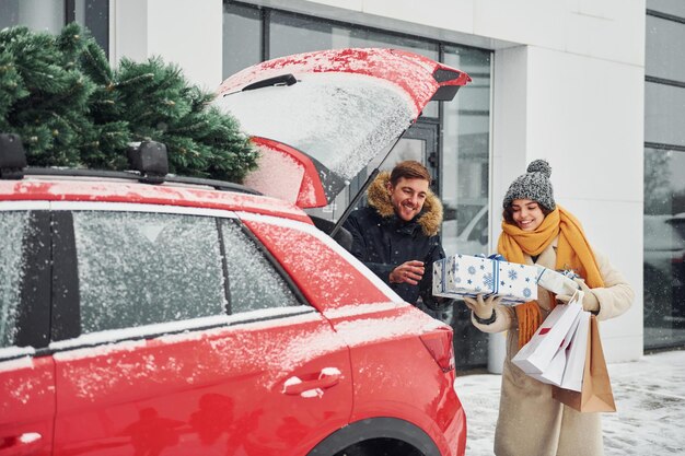 Un jeune couple avec des coffrets cadeaux est près d'une voiture avec un arbre sur le dessus. Ensemble à l'extérieur à l'heure d'hiver.