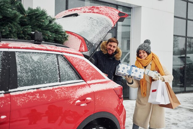 Un jeune couple avec des coffrets cadeaux est près d'une voiture avec un arbre sur le dessus. Ensemble à l'extérieur à l'heure d'hiver.