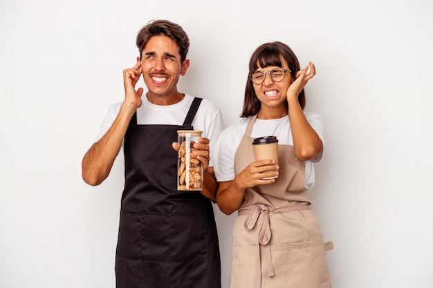 Jeune couple de commis de magasin de race mixte isolé sur fond blanc couvrant les oreilles avec les mains.