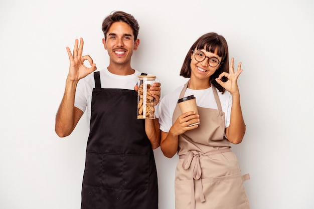 Jeune couple de commis de magasin de race mixte isolé sur fond blanc gai et confiant montrant un geste ok.