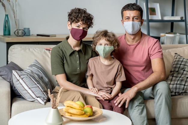Jeune couple contemporain et leur petit fils dans des masques de protection assis sur un canapé dans le salon tout en restant à la maison pour la quarantaine