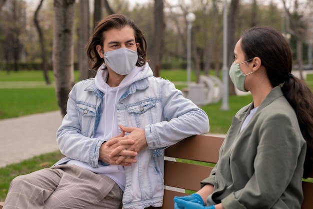 Jeune couple contemporain en vêtements décontractés et masques de protection assis sur un banc en bois