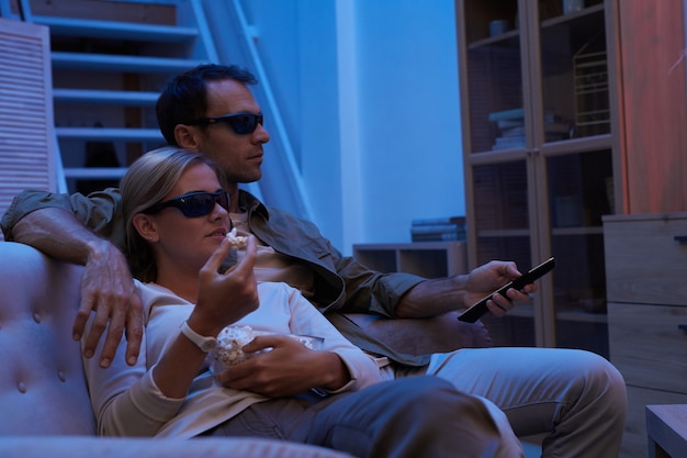 Photo jeune couple dans des lunettes 3d de manger du pop-corn assis sur un canapé et regarder un film 3d à la maison