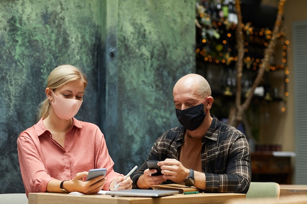 Jeune couple dans des masques de protection à l'aide de téléphones mobiles pendant le déjeuner au café