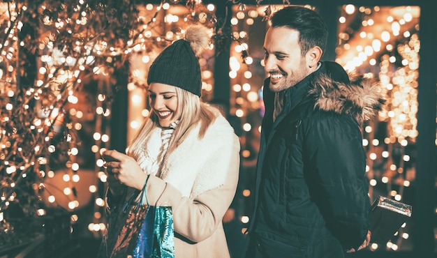 Jeune couple debout dans un centre commercial avec vitrine lumineuse en arrière-plan. Une belle femme s'amusant en regardant un smartphone.