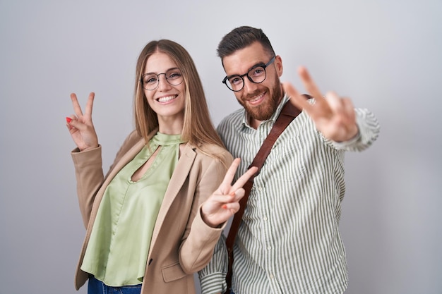 Jeune couple debout sur fond blanc souriant regardant la caméra montrant les doigts faisant le signe de la victoire. numéro deux.