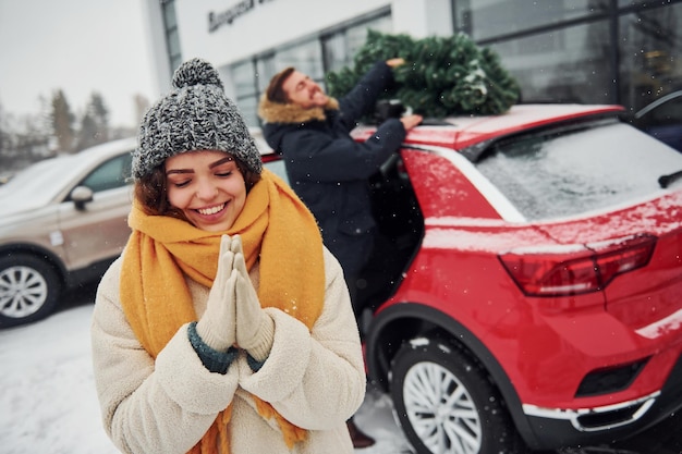 Jeune couple debout près de la voiture avec un arbre sur le dessus. Ensemble à l'extérieur à l'heure d'hiver.