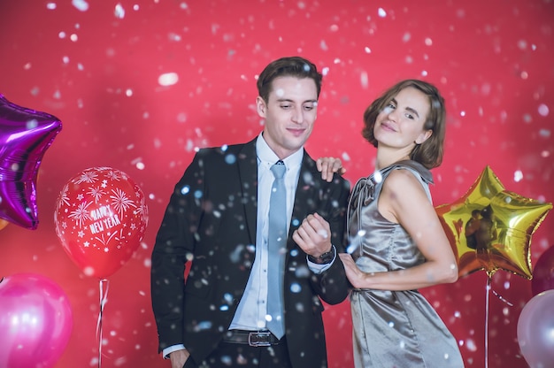 Le jeune couple est heureux parmi les confettis qui tombent devant. Il y a des ballons colorés à côté d'eux et le mur rouge dans le concept du nouvel an et du jour de Noël.