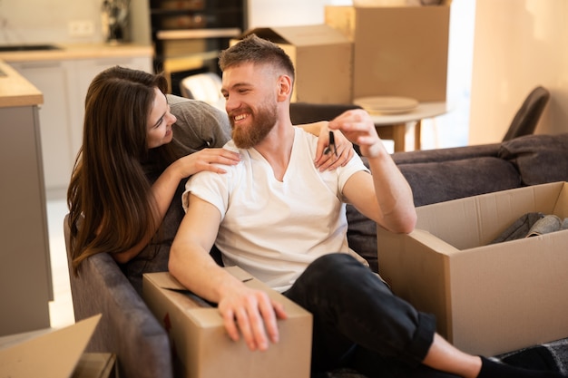 Jeune couple européen heureux célébrant à la nouvelle maison. Homme assis sur le canapé et tenant la clé. Boîtes en carton avec des choses. Concept de déménagement dans un nouvel appartement. Idée de jeune famille. Intérieur du studio