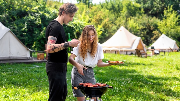 Jeune couple faisant frire de la viande sur le gril et buvant de la bière. Verdure autour. Glamping