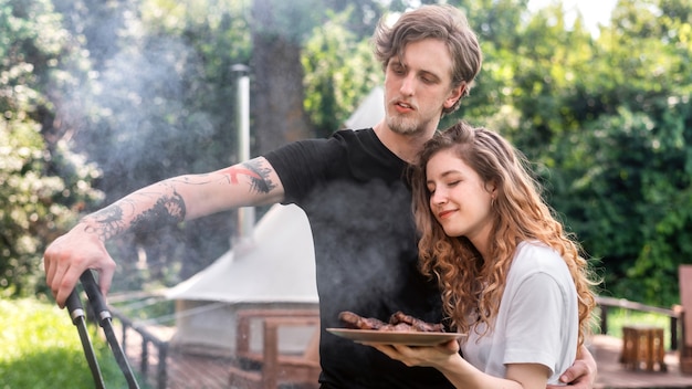 Jeune couple faisant frire de la viande sur le gril et la mettant sur l'assiette. Verdure autour. Glamping