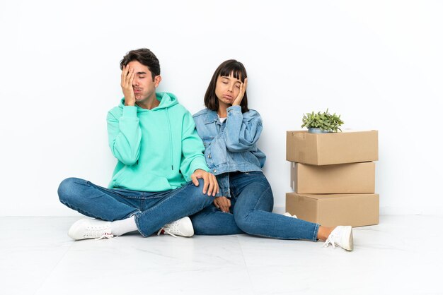 Jeune couple faisant un geste tout en ramassant une boîte pleine de choses assis sur le sol isolé sur fond blanc avec une expression faciale surprise et choquée