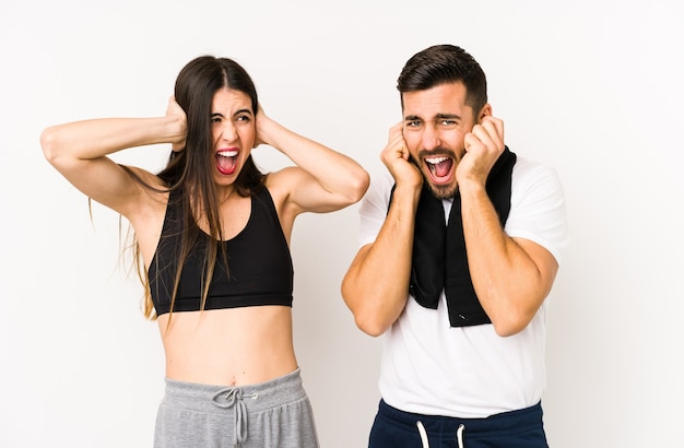 Jeune couple de fitness caucasien isolé couvrant les oreilles avec les mains en essayant de ne pas entendre un son trop fort.