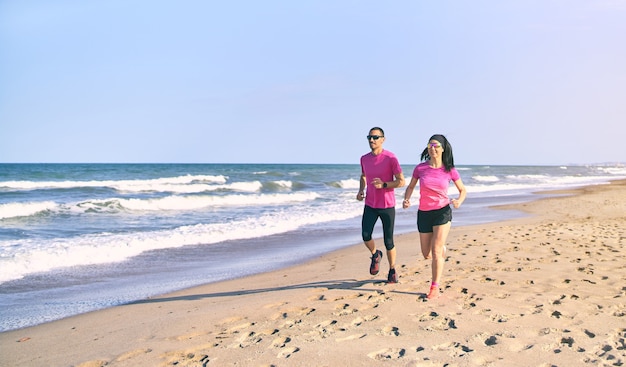 Jeune couple en forme qui court sur la plage au lever du soleil. Début de journée sain. Porter des vêtements de sport roses et noirs. Courir au bord de la mer.