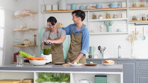 Jeune couple gay souriant faisant du jus sain avec une machine à jus dans la cuisine à la maison LGBTQ et concept de diversité