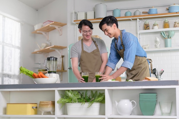 Jeune couple gay souriant faisant du jus sain avec une machine à jus dans la cuisine à la maison