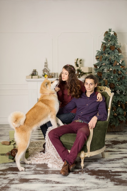 Jeune couple heureux câlins adorable chien akita inu tout en étant assis sur un élégant fauteuil rétro pour les vacances de Noël à la maison