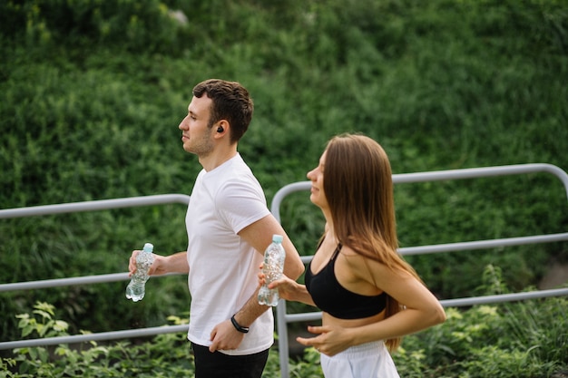 Jeune couple heureux courant dans le parc de la ville avec une bouteille d'eau dans les mains, sports communs, gaieté, mode de vie sain pour le sport en ville, remise en forme