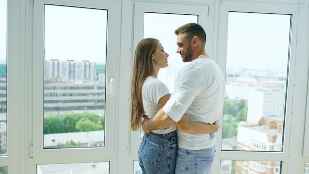 Jeune couple heureux embrassant debout près de la fenêtre et profitant de la vue depuis le nouvel appartement