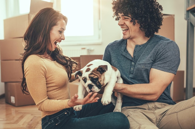 Le jeune couple heureux emménage dans une nouvelle maison. Ils tiennent leur petit chiot après avoir apporté des boîtes avec des choses dans leur nouvelle maison.