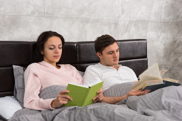 Jeune couple heureux lisant des livres en position couchée dans le lit et portant un pyjama dans la chambre de style loft avec des couleurs grises