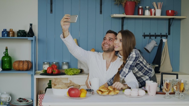 Jeune couple heureux prenant un selfie tout en prenant son petit déjeuner dans la cuisine à la maison