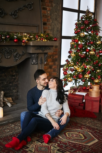 Le jeune couple heureux, en pulls chauds et chaussettes rouges, étreignant et assis sur le tapis authentique près de la cheminée et du sapin de Noël. Célébration de Noël et vacances du nouvel an. Ambiance de Noël.