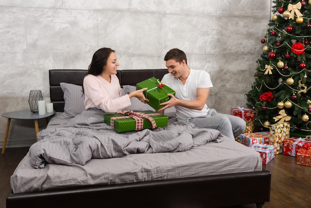 Jeune couple heureux en pyjama se donnant des cadeaux assis sur le lit dans la chambre de style loft le matin de Noël