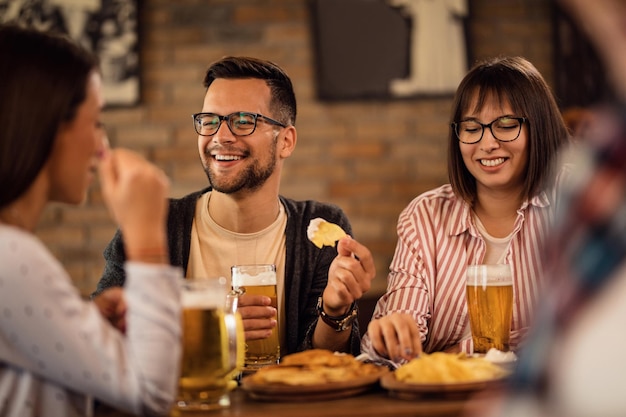 Jeune couple heureux s'amusant avec des amis tout en buvant de la bière et en prenant une collation dans une taverne