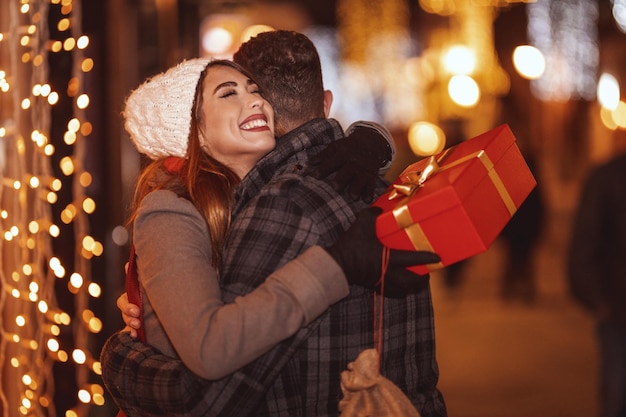 Un jeune couple heureux se promène le long de la rue de la ville avec des vacances lumineuses en arrière-plan. Un homme souriant donne un cadeau à sa petite amie heureuse et ils s'embrassent dans une nuit de Noël.