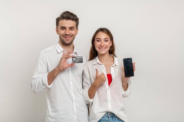 Jeune couple heureux tenir une carte bancaire de crédit à l'aide d'un smartphone avec le pouce vers le haut isolé sur fond blanc