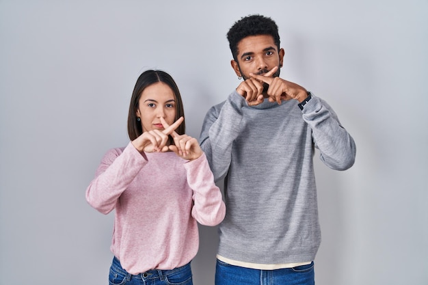 Jeune couple hispanique debout ensemble expression de rejet croisant les doigts faisant signe négatif