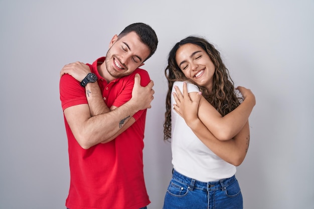 Jeune couple hispanique debout sur fond isolé se serrant dans ses bras heureux et positif souriant confiant amour de soi et soins personnels