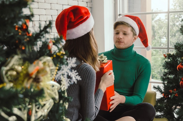 Jeune couple homme avec Bonnet de Noel rouge surprenant et donnant un coffret cadeau de Noël à une copine à Noël dans la maison