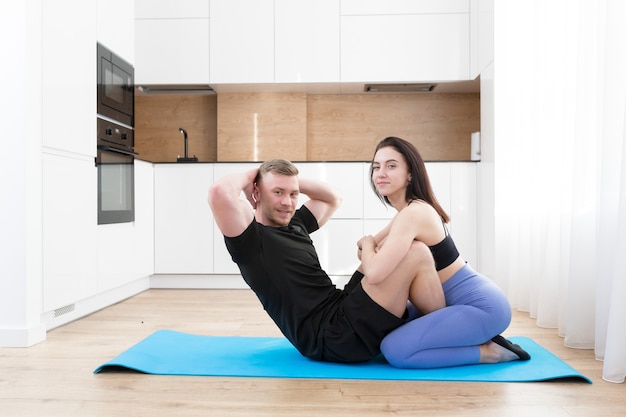 Jeune couple homme et femme faisant du fitness ensemble à la maison sur le sol, deux personnes faisant des exercices de sport dans la cuisine sur un tapis de sport