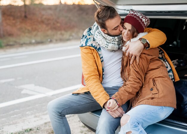 Jeune couple d'homme et femme, portant des vêtements chauds, assis sur le coffre de la voiture, étreignant un jour d'hiver