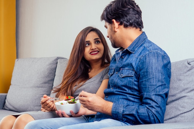 Jeune couple indien assis dans leur salon et mangeant une salade de légumes.