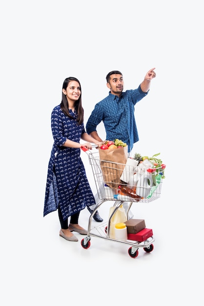 Jeune couple indien avec caddie ou chariot plein d'épicerie, de légumes et de fruits. Photo pleine longueur isolée sur mur blanc