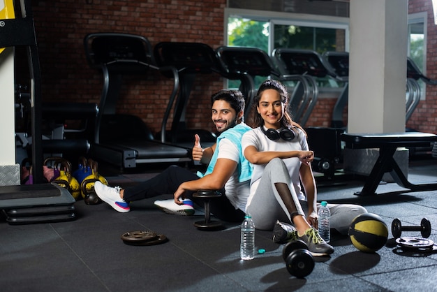 Jeune couple indien dans une salle de sport se reposant ou faisant une pause dans l'exercice, buvant de l'eau, montrant des expressions positives