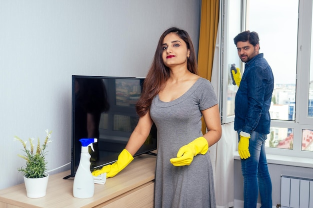 Jeune couple indien emménageant dans une nouvelle maison, nettoyant et déballant une grande fenêtre avec un fond de rideau jaune élégant.