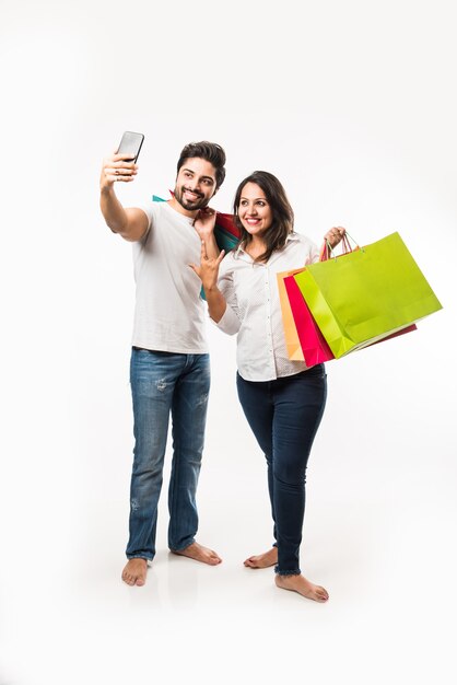 Jeune couple indien sacs à provisions et téléphone intelligent ou mobile, en cliquant sur selfie ou en localisant le magasin, debout isolé sur fond blanc. mise au point sélective