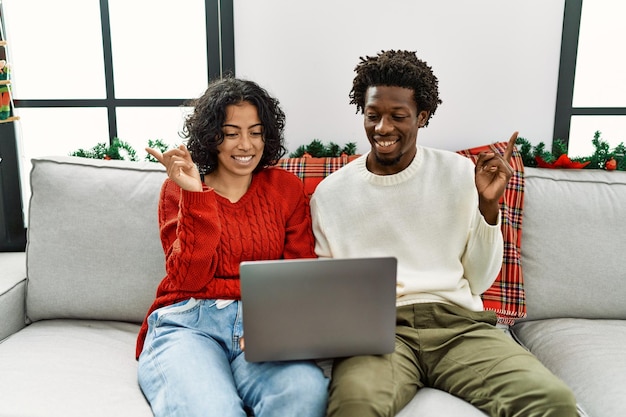 Jeune couple interracial assis sur le canapé à noël à l'aide d'un ordinateur portable souriant heureux pointant avec la main et le doigt sur le côté