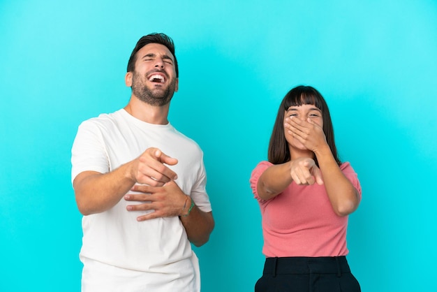Jeune couple isolé sur fond bleu pointant du doigt quelqu'un et riant beaucoup