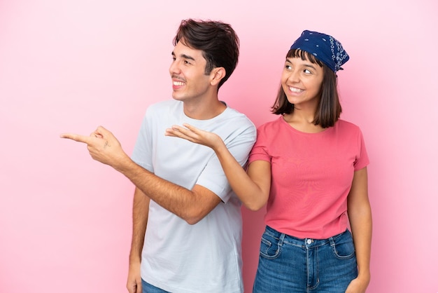 Jeune couple isolé sur fond rose pointant vers l'arrière et présentant un produit