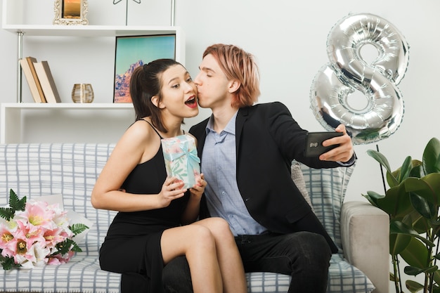 Jeune couple le jour de la femme heureuse tenant présent prendre un selfie assis sur un canapé dans le salon