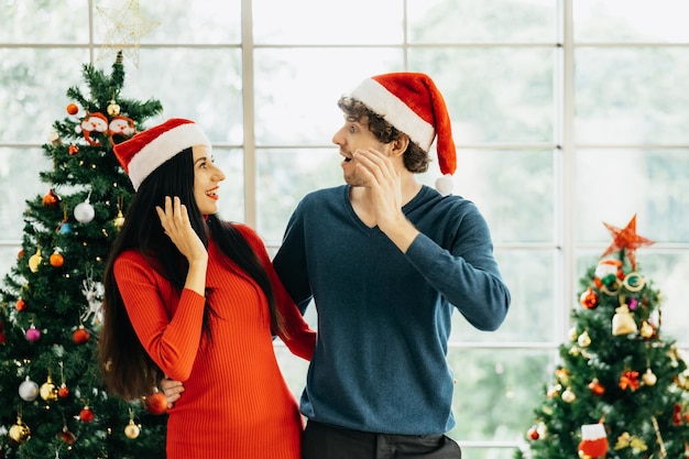 Jeune couple joyeux en chapeaux de Père Noël à l'aide d'une tablette assis entouré de coffrets cadeaux colorés profitant de la célébration de Noël à la maison.