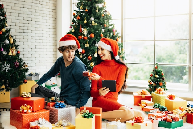 Jeune couple joyeux en chapeaux de Père Noël à l'aide d'une tablette assis entouré de coffrets cadeaux colorés profitant de la célébration de Noël à la maison.