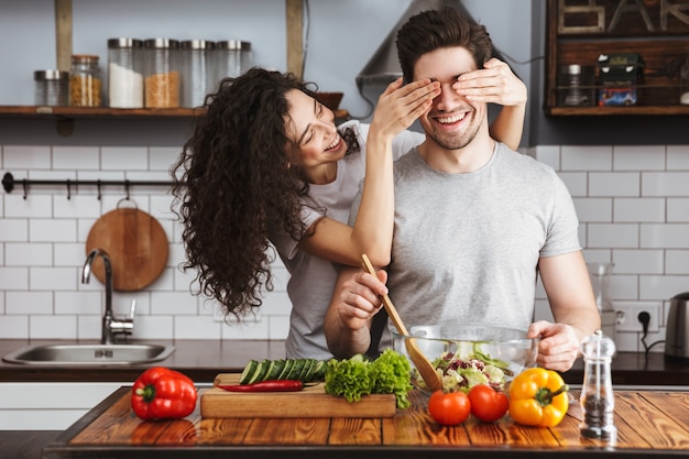 Jeune couple joyeux et excité cuisinant une salade saine assis à la cuisine, la femme couvre les yeux des hommes