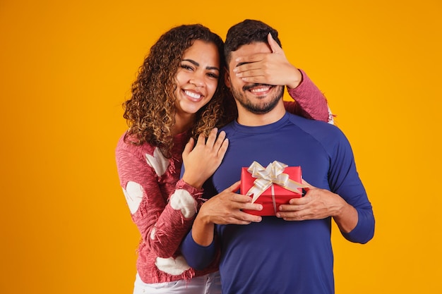 Jeune couple joyeux et excité debout isolé sur un fond jaune couvrant leurs yeux surpris tenant un cadeau