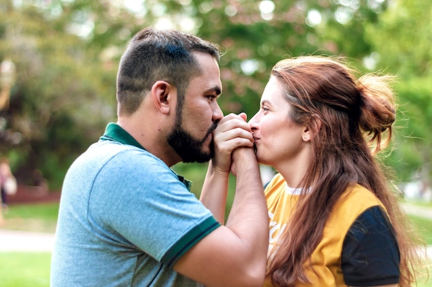 Jeune couple latin formé d'une fille aux cheveux roux et d'un garçon aux cheveux courts et à la barbe, se font face, les mains entrelacées au niveau des lèvres et se regardent dans les yeux.